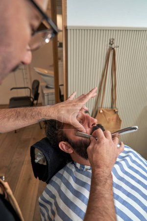 Foto de Peluquero serio irreconocible en anteojos barba de corte de cliente masculino con hoja recta en la barbería moderna - Imagen libre de derechos