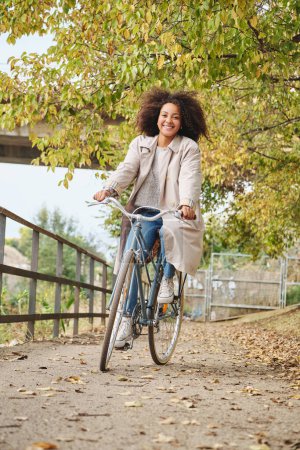 Téléchargez les photos : Corps complet de femme afro-américaine avec les cheveux bouclés dans des vêtements décontractés à vélo sur le chemin pendant la journée d'automne - en image libre de droit