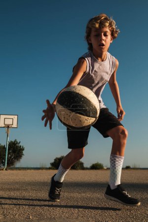 Téléchargez les photos : Corps entier de l'enfant en vêtements de sport lancer le ballon tout en jouant au streetball sur le terrain de sport contre ciel bleu sans nuages pendant l'entraînement - en image libre de droit