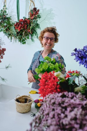 Photo for Positive adult female florist in glasses and apron standing near counter with bouquet of green trachelium surrounded by blooming flowers in floral shop - Royalty Free Image