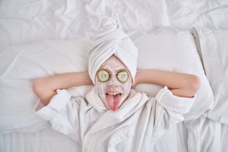 Photo for Top view of adorable positive kid with towel on head and facial mask with cucumbers relaxing during skincare procedure and showing tongue - Royalty Free Image