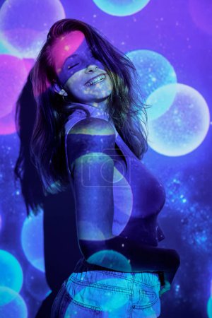 Foto de Vista lateral de la joven optimista con el pelo largo sonriendo alegremente con los ojos cerrados y tocando el vientre mientras baila bajo la proyección de destellos de colores - Imagen libre de derechos