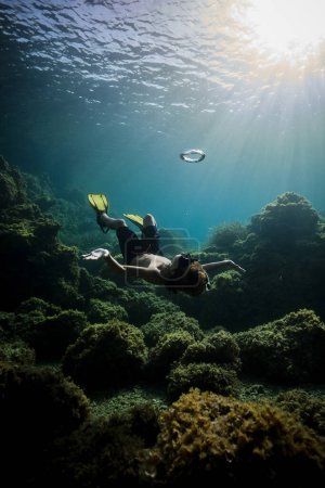 Foto de Cuerpo completo de buceador irreconocible en máscara de snorkel y aletas nadando bajo el agua de mar azul cerca del arrecife de coral y mirando al sol - Imagen libre de derechos