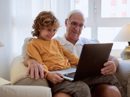 Foto de Sonriendo hombre mayor con nieto viendo interesante vídeo en el ordenador portátil mientras están sentados juntos en el sillón en la sala de estar en casa - Imagen libre de derechos