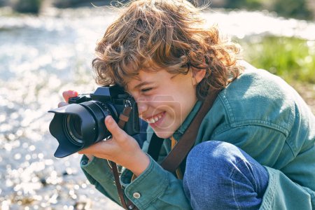 Photo for Side view of happy boy taking picture with professional photo camera while sitting on haunches near flowing river on blurred background - Royalty Free Image