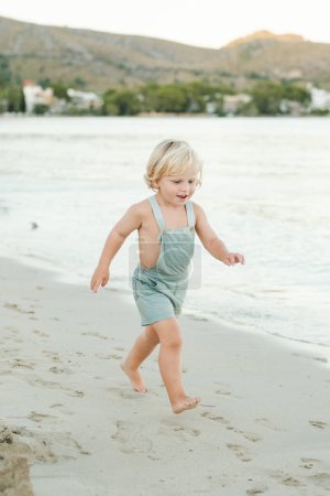 Téléchargez les photos : Pleine longueur de blond pieds nus bambin dans l'ensemble courir sur le rivage sablonneux contre la mer sereine tout en profitant de l'été - en image libre de droit