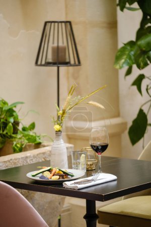 Foto de Mesa de madera con ramo de flores y plato con copa de vino tinto servido para la cena en el restaurante - Imagen libre de derechos