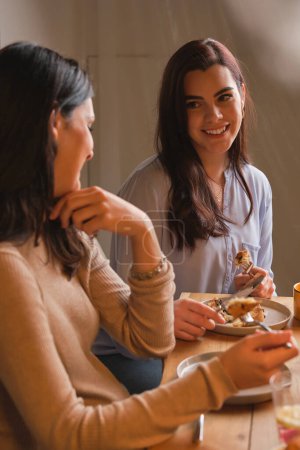Téléchargez les photos : Des jeunes femmes souriantes en vêtements décontractés mangeant ensemble de délicieux gâteaux faits maison et parlant dans la cuisine à la maison - en image libre de droit