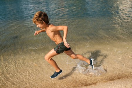 Téléchargez les photos : Full body side view of happy shirtless preteen boy enjoy summer holidays while having fun and running along sandy shore in clear sea water - en image libre de droit
