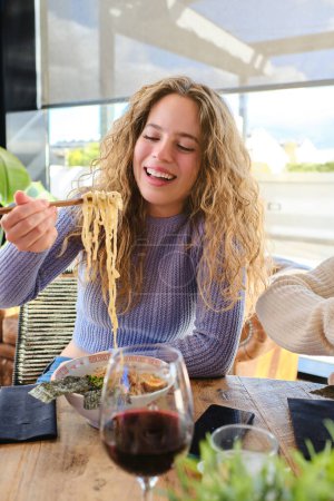 Photo for Young blond female smiling and taking noodles with chopsticks while sitting at table in front of glass of red wine - Royalty Free Image