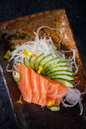 Photo for Top view of raw salmon fillet decorated with cucumber slices flowers and shredded daikon placed on marble tray against black background - Royalty Free Image