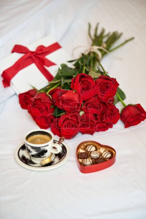 Foto de De arriba de la composición del regalo romántico que consiste en el ramo de las rosas rojas la taza del café fresco y la caja en forma de corazón de los bombones puestos de la cama en la habitación del hotel - Imagen libre de derechos