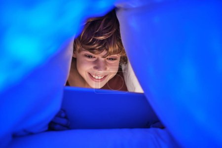 Foto de Niño feliz viendo vídeo en la tableta con sonrisa mientras está acostado debajo de la manta en el dormitorio con poca luz en casa por la noche - Imagen libre de derechos