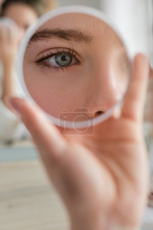 Photo for Eye of crop unrecognizable female reflecting in small round mirror in hand while sitting at table during daily routine at home - Royalty Free Image