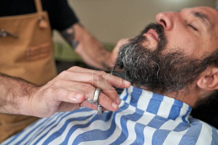 Téléchargez les photos : Barbier anonyme à l'aide de ciseaux pour couper les cheveux du client masculin barbu tout en travaillant dans le salon de coiffure - en image libre de droit