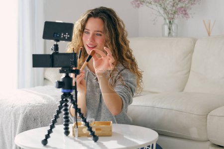 Téléchargez les photos : Jeune femme vlogger avec de longs cheveux ondulés montrant ensemble de brosses pour le maquillage du visage à la caméra dans le salon à la maison - en image libre de droit
