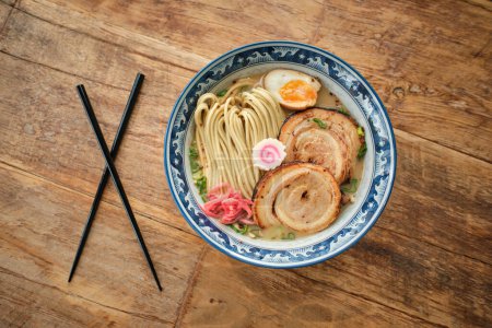Téléchargez les photos : Vue du dessus de la soupe de ramen appétissante aux nouilles aux œufs durs et à la viande servie dans un bol en céramique placé près des baguettes sur une table en bois - en image libre de droit