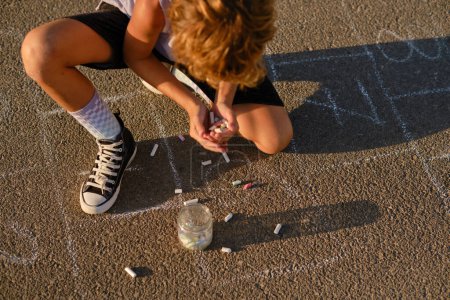 Photo for From above of unrecognizable preteen child with pile of colorful pieces of chalks sitting on asphalt ground with hopscotch in summer - Royalty Free Image