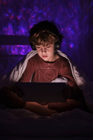 Photo for Concentrated boy surfing modern tablet while sitting under blanket in dark bedroom with dim light at night time in apartment - Royalty Free Image