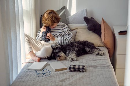 Photo for Schoolchild with old photo camera sitting with crossed legs near purebred dog on bed at home - Royalty Free Image