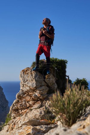 Photo for Full length of male traveler with rucksack standing on rough stony edge of mountain and observing picturesque scenery while looking away - Royalty Free Image