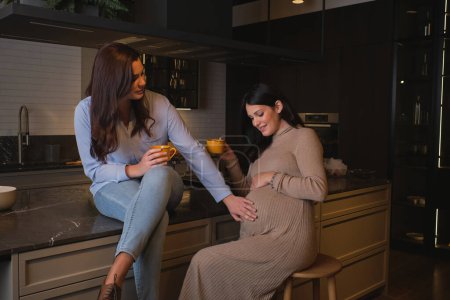 Téléchargez les photos : Femme positive avec tasse touchant le ventre d'une amie enceinte et souriant tout en étant assis sur une table en marbre dans une cuisine confortable à la maison le week-end - en image libre de droit