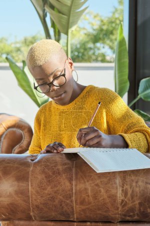 Téléchargez les photos : Une jeune afro-américaine concentrée écrit des notes dans un journal assis sur un canapé à la maison - en image libre de droit