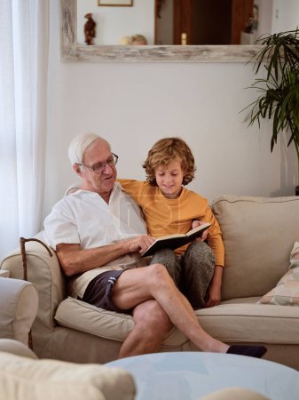 Foto de Hombre de pelo gris senior en ropa casual sentado en el sofá y libro de lectura con nieto en la acogedora sala de estar - Imagen libre de derechos