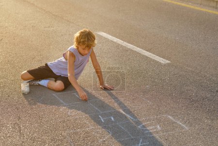 Photo for High angle of peaceful preteen child sitting on roadway and drawing hopscotch with chalk while entertaining at weekend in summer - Royalty Free Image