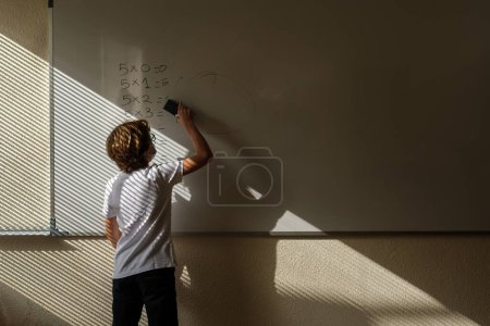 Photo for Back view of faceless schoolboy erasing answers of mathematical examples while standing near whiteboard in classroom during lesson - Royalty Free Image