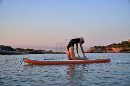 Téléchargez les photos : Corps complet de femme sportive en maillot de bain à manches longues prenant la pose d'Ushtrasana tout en faisant du yoga sur paddleboard en mer bleue au coucher du soleil - en image libre de droit