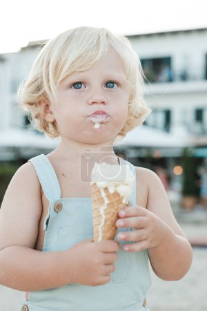 Photo for Fair haired child eating ice cream cone while standing on blurred street with smeared mouth in daylight - Royalty Free Image
