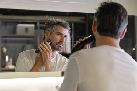 Photo for Concentrated male in casual clothes shaving beard using professional trimmer while looking at reflection in mirror at home - Royalty Free Image