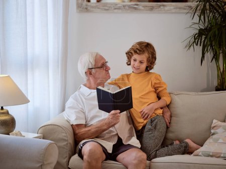 Téléchargez les photos : Grand-père attentionné avec mignon petit-fils en tenue décontractée lecture livre intéressant tout en étant assis sur le canapé dans le salon à la maison - en image libre de droit