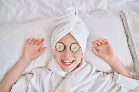 Foto de Niño con máscara de pepino con turbante toalla y albornoz relajante en la cama y sonriendo durante los procedimientos de belleza en el dormitorio en casa - Imagen libre de derechos