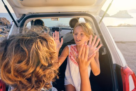 Photo for Happy siblings in summer outfits playing patty cake game near opened car trunk during vacation on sandy seashore - Royalty Free Image
