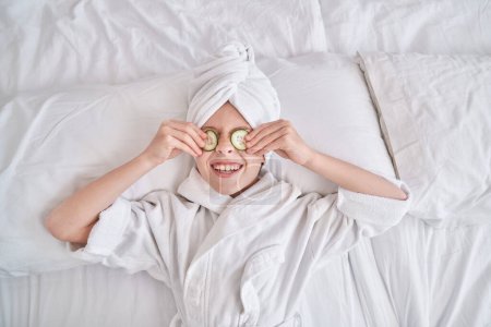 Foto de Vista superior del niño sonriente con turbante de toalla y albornoz poniendo rodajas de pepino en los ojos y divirtiéndose durante los procedimientos de spa en casa - Imagen libre de derechos