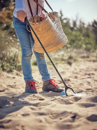 Téléchargez les photos : Vue latérale de la culture personne méconnaissable avec sac en osier et poubelle ramassage bouteille en plastique sur la plage de sable - en image libre de droit
