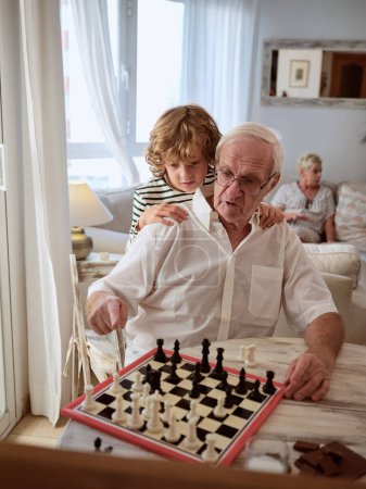 Téléchargez les photos : Focalisé préadolescent garçon étreignant grand-père âgé tout en apprenant à jouer aux échecs à table dans le salon à la maison - en image libre de droit