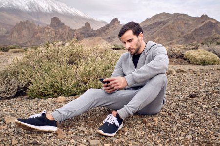 Photo for Side view of bearded concentrated male in activewear texting message on mobile phone while resting on stony ground near volcano Teide in Tenerife in Canary Islands in Spain - Royalty Free Image
