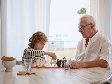 Téléchargez les photos : Focalisé garçon et homme âgé dans des lunettes assis à la table en bois avec des cookies et jeu de société dans la cuisine à la maison - en image libre de droit