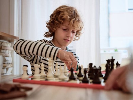 Photo for Portrait of focused child sitting at wooden table and learning to play chess with crop person at table in kitchen at home - Royalty Free Image