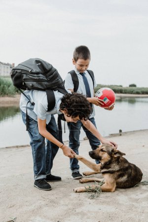 Photo for Side view of male pupils in school uniform with backpack holding ball while standing with obedient dog on shore of river in daylight - Royalty Free Image
