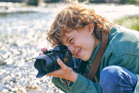 Téléchargez les photos : Vue latérale d'un garçon optimiste prenant des photos avec un appareil photo assis sur le rivage près d'une rivière qui coule sur fond flou - en image libre de droit