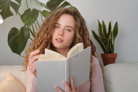 Téléchargez les photos : Jeune femme réfléchie avec de longs cheveux bouclés lecture roman intéressant et assis sur un canapé blanc dans le salon moderne - en image libre de droit