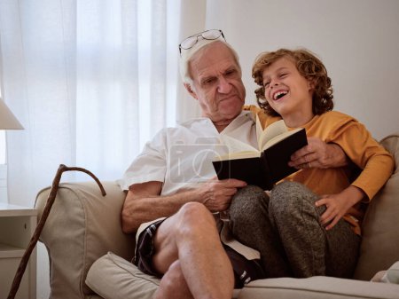 Téléchargez les photos : Garçon souriant étreignant homme âgé tenant le livre et assis sur le canapé confortable le jour de congé dans le salon - en image libre de droit