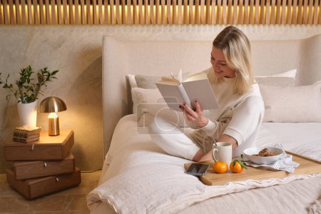 Téléchargez les photos : Corps complet de femme souriante en vêtements de nuit blancs assis sur un lit doux près d'un plateau en bois avec petit déjeuner et lecture livre intéressant dans la chambre à coucher à la maison - en image libre de droit