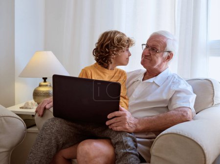 Téléchargez les photos : Petit-fils et grand-père positifs avec netbook se regardant tout en étant assis ensemble dans un fauteuil confortable dans le salon léger - en image libre de droit