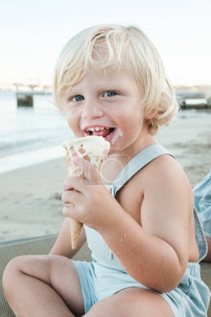 Photo for Adorable blond haired toddler in overall sitting on rug and eating yummy ice cream while looking at camera during weekend on beach - Royalty Free Image