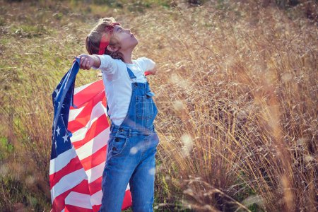 Téléchargez les photos : Vue latérale du contenu kid en jeans debout dans le champ jaune avec drapeau américain dans les bras écartés - en image libre de droit